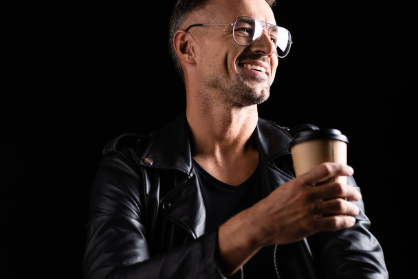 Selective focus of smiling handsome man in sunglasses holding coffee to go isolated on black  - Φωτογραφία, εικόνα