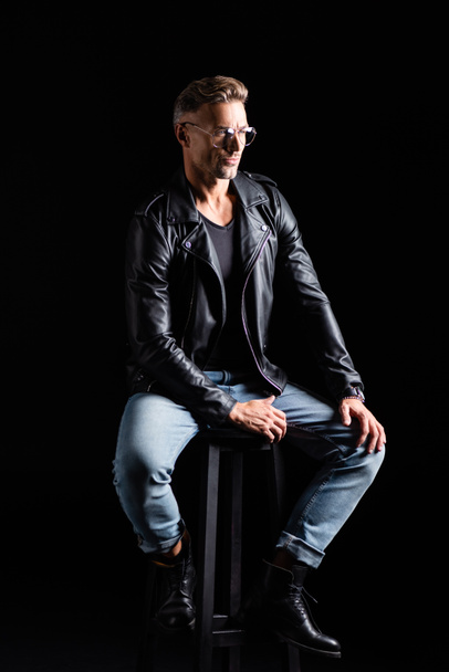 Comprimento total de homem elegante em jeans e jaqueta de couro sentado na cadeira isolada em preto
  - Foto, Imagem