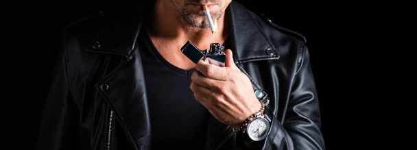 Vista recortada del hombre en la chaqueta de cuero iluminación cigarrillo con encendedor aislado en negro, tiro panorámico
 - Foto, imagen