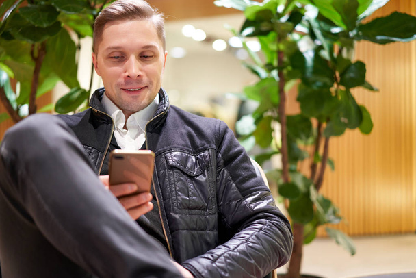 Young man in leather jacket with smartphone in his hands sitting on chair in store against green plant - Foto, Bild