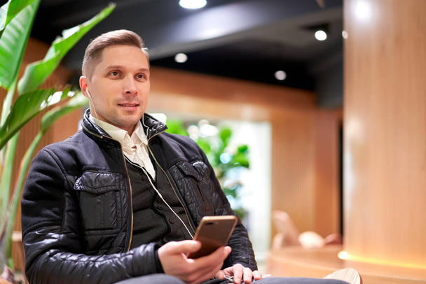Brrunet man with smartphone looking at camera sitting on chair in store against green plant - Фото, изображение