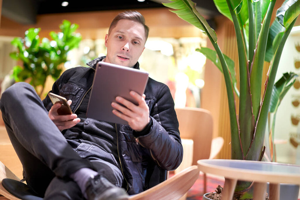 Young man holding phone and tablet in his hands sitting on chair in store against green plant - Photo, Image