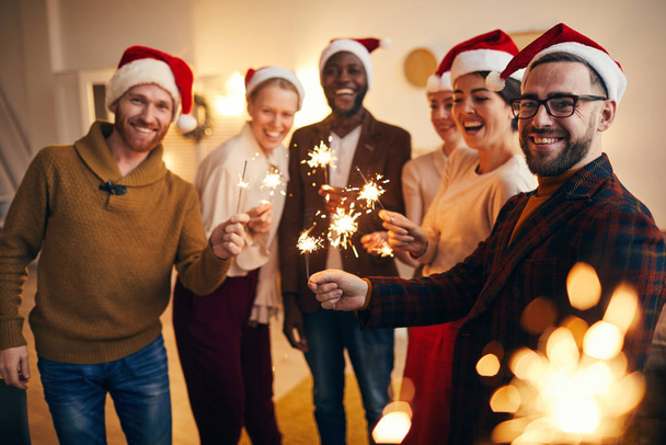 Monietninen ryhmä ihmisiä, joilla on kuohuvaloja ja jotka katsovat kameraa nauttien joulujuhlista ystävien ja perheen kanssa
 - Valokuva, kuva