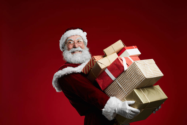 Waist up portrait of smiling Santa Claus holding stack of Christmas presents over red background, copy space - Photo, Image