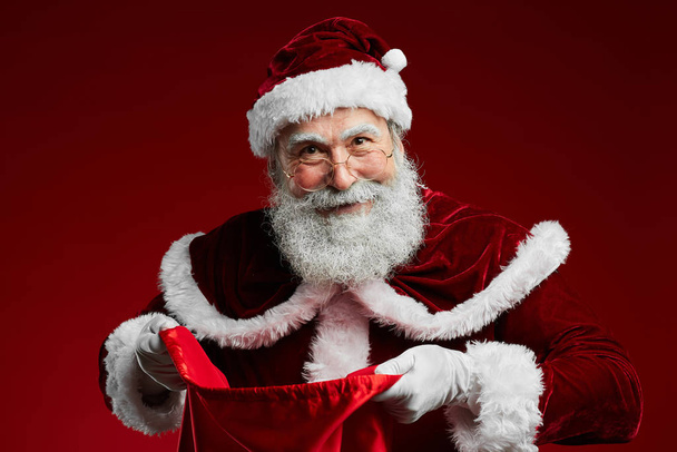 Taille nach oben Porträt eines lächelnden Weihnachtsmannes, der einen Sack mit Weihnachtsgeschenken in der Hand hält, während er über rotem Hintergrund steht, Kopierraum - Foto, Bild