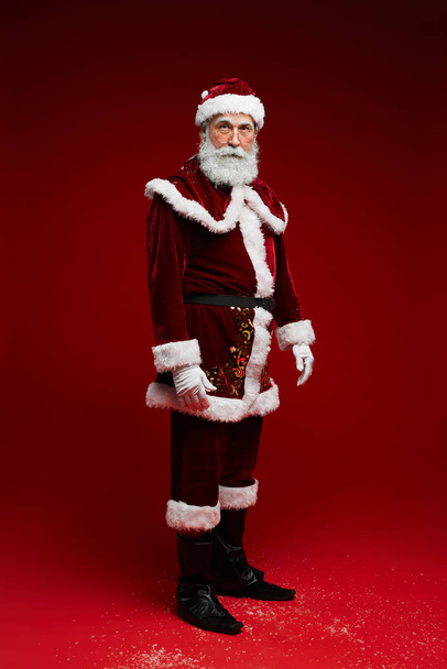 Full length portrait of classic Santa Claus looking at camera while posing against red background in studio, copy space - Zdjęcie, obraz