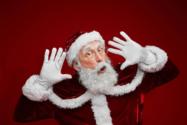 Waist up portrait of surprised Santa Claus looking at camera while posing against red background in studio, copy space - Photo, Image