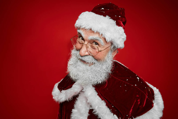 Gülümseyen Noel Baba 'nın portresi kameraya yaramazca bakarken stüdyoda kırmızı arka planda poz veriyor, boşluğu kopyalıyor. - Fotoğraf, Görsel