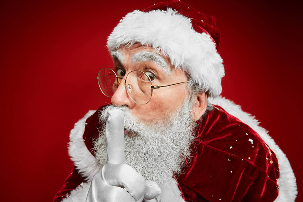 Detail portrét klasické Santa Claus uvedení prst do úst vyprávění tajemství, zatímco stojí na červeném pozadí - Fotografie, Obrázek
