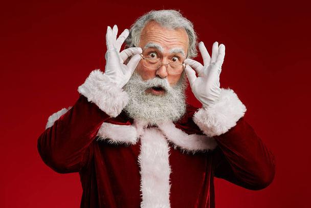 Taillenhoch Porträt des überraschten Weihnachtsmannes, der in die Kamera schaut und die Brille anpasst, während er vor rotem Hintergrund im Studio posiert, Kopierraum - Foto, Bild