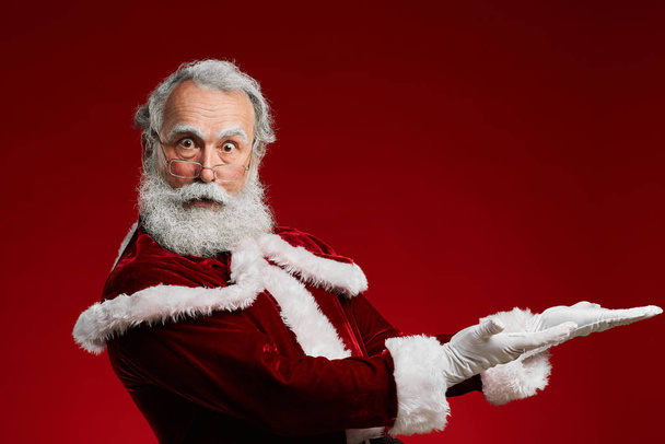 Cintura até retrato de Papai Noel surpreso olhando para a câmera e apontando ao posar contra fundo vermelho, espaço de cópia
 - Foto, Imagem