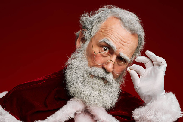 Retrato de cabeza y hombros del sospechoso Santa Claus ajustando las gafas mientras mira la cámara sobre fondo rojo, espacio para copiar
 - Foto, Imagen