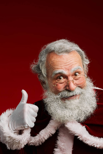 Retrato de Santa Claus sonriente mostrando los pulgares hacia arriba mientras posando sobre fondo rojo en el estudio, espacio de copia
 - Foto, Imagen