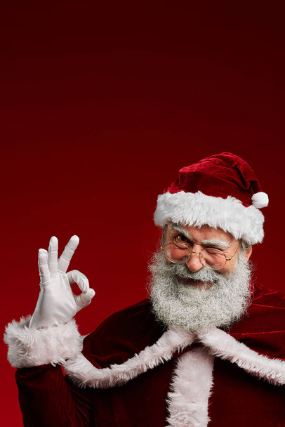 Portrait taille haute de clin d'oeil Père Noël regardant la caméra et montrant OK signe tout en posant sur fond rouge, copier l'espace
 - Photo, image