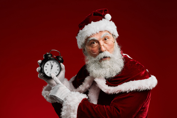 Retrato de la cintura hacia arriba de Santa Claus clásico apuntando al reloj mientras posando sobre fondo rojo en el estudio, espacio de copia
 - Foto, Imagen