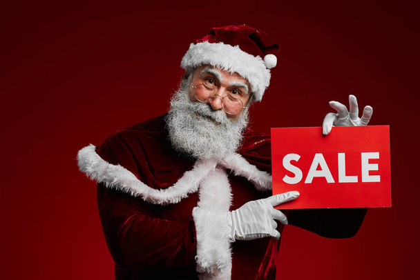 Waist up portrait of smiling Santa Claus holding SALE sign and looking at camera while standing against red background, copy space - Фото, изображение