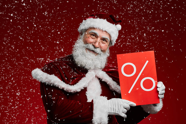 Cintura hacia arriba retrato de Santa Claus sonriente sosteniendo Venta signo y mirando a la cámara mientras posando sobre fondo rojo con nieve cayendo, espacio de copia
 - Foto, imagen