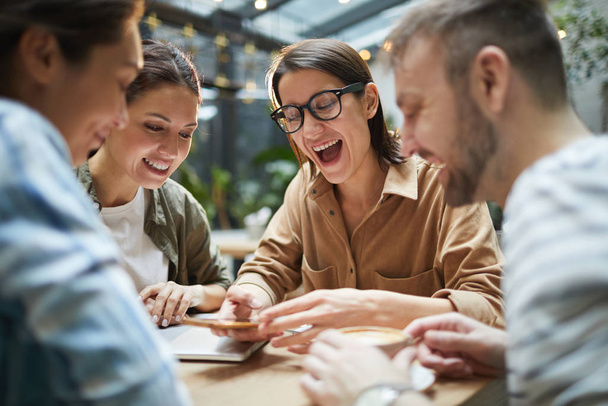Groep hedendaagse jongeren die naar het smartphone scherm kijken en vrolijk lachen terwijl ze samen lunchen in café - Foto, afbeelding