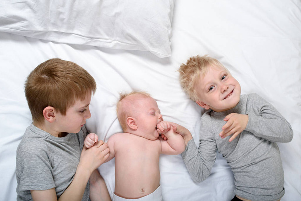 Dva starší bratři si hrají a smějí se s nejmladším dítětem v bílé posteli. Šťastné dětství, velká rodina - Fotografie, Obrázek