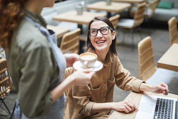 Retrato de alto ángulo de la joven mujer sonriendo a la camarera mientras trabaja en la mesa de la cafetería, espacio de copia
 - Foto, Imagen