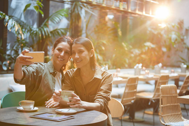 Portrait of two contemporary women taking selfie photo while enjoying meeting in sunlit care terrace, copy space - 写真・画像