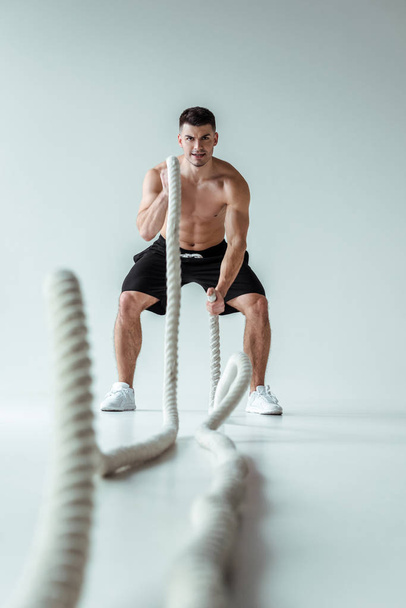 selective focus of sexy muscular bodybuilder with bare torso exercising with battle rope on grey background - Photo, Image