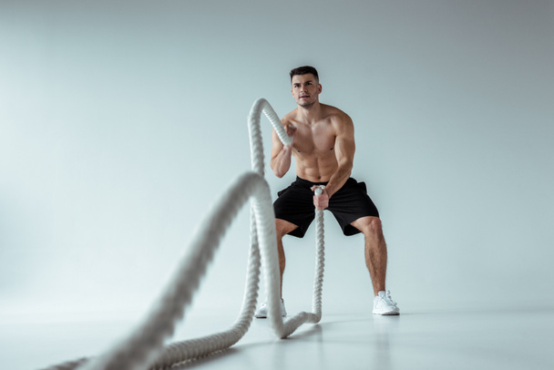 selective focus of sexy muscular bodybuilder with bare torso exercising with battle rope on grey background - Foto, afbeelding
