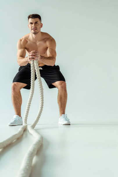 selective focus of sexy muscular bodybuilder with bare torso exercising with battle rope on grey background - Foto, Imagem