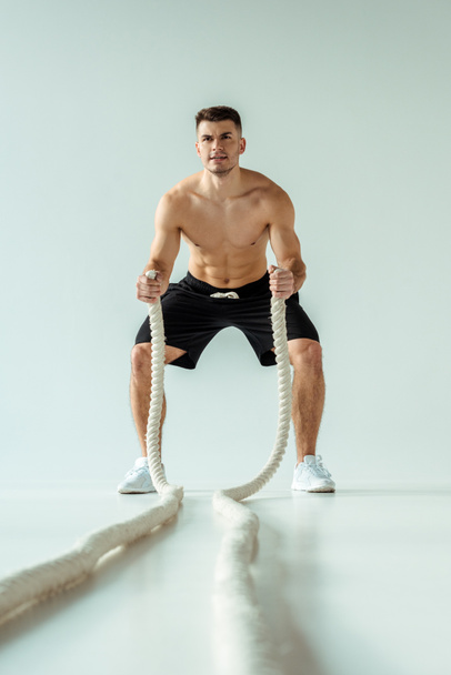 selective focus of sexy muscular bodybuilder with bare torso exercising with battle rope on grey background - Foto, immagini