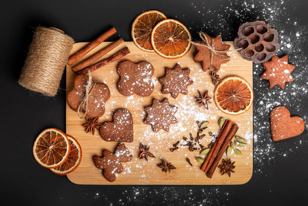 Gingerbread cookies with spices on a wooden plank against the black background. Cinnamon sticks, orange slices, star anise, clove, cardamom and powdered sugar in a beautiful composition. Holiday atmosphere. Cookies flavored with ginger. - Fotografie, Obrázek