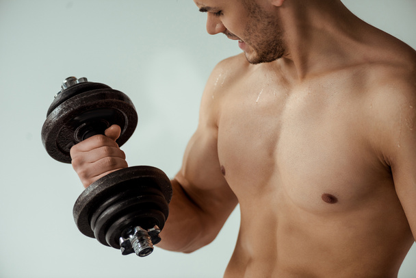sweaty tense muscular bodybuilder with bare torso working out with dumbbell isolated on grey - Foto, Imagen
