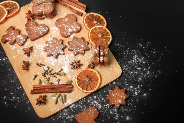 Gingerbread cookies with spices on a wooden plank against the black background. Cinnamon sticks, orange slices, star anise, clove, cardamom and powdered sugar in a beautiful composition. Holiday atmosphere. Cookies flavored with ginger. - Фото, изображение