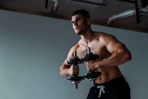 low angle view of tense muscular bodybuilder with bare torso working out with dumbbells - Zdjęcie, obraz