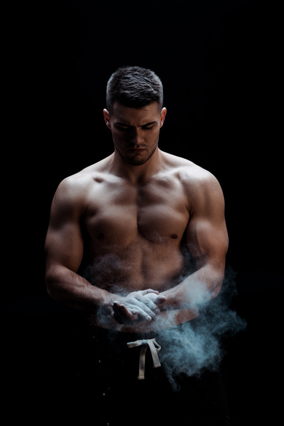 sexy muscular bodybuilder with bare torso applying talcum powder on hands on black background with smoke  - Photo, Image