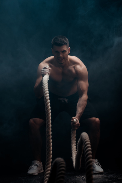sexy muscular bodybuilder with bare torso excising with battle rope on black background with smoke  - Foto, imagen