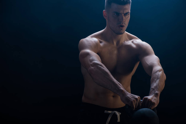 tense sexy muscular bodybuilder with bare torso excising with kettlebell on black background - Foto, Imagem