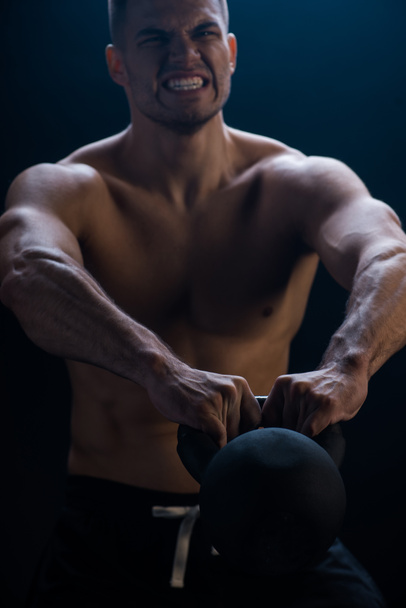tense sexy muscular bodybuilder with bare torso excising with kettlebell on black background - Photo, Image