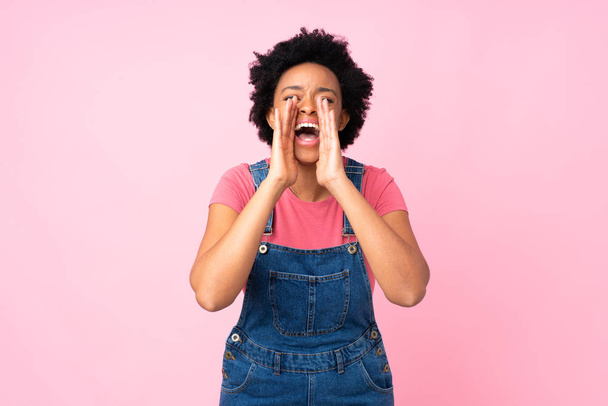 African american woman with overalls over isolated pink background shouting with mouth wide open - Fotografie, Obrázek