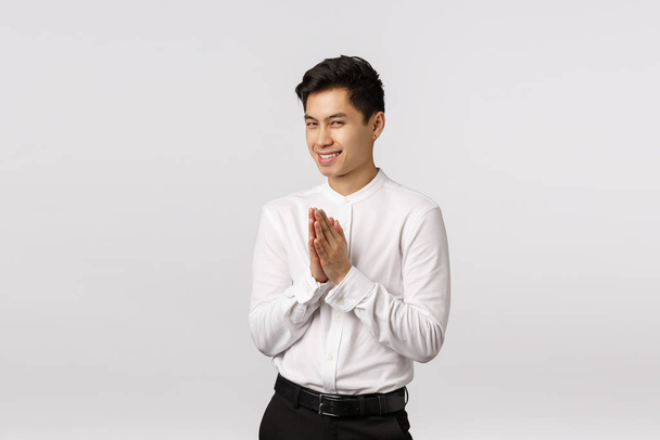 Hideous, astuto bonito jovem empresário asiático tem algo mal ou gênio em mente, esfregue palmas perto do peito, squinting misterioso e sorrir, tem plano perfeito, de pé fundo branco
 - Foto, Imagem