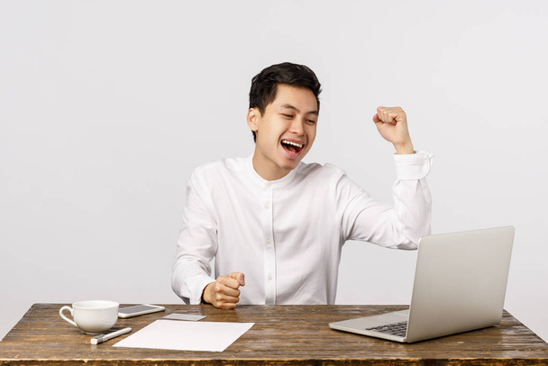Happy, rejoicing young asian male entrepreneur fist pump, raising hand in victory, celebration gesture, sitting desk with documents at work, looking laptop screen, smiling telling yes - Foto, immagini