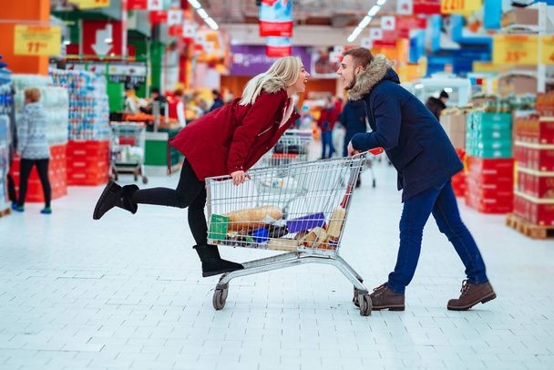 junger hübscher Kerl reitet ein Mädchen in einem Supermarkt in einem Einkaufswagen - Foto, Bild