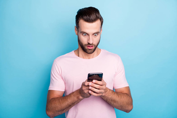 Photo of attractive guy holding telephone hands open mouth reading bad news negative post comments followers wear casual pink t-shirt isolated blue color background - Foto, Imagem