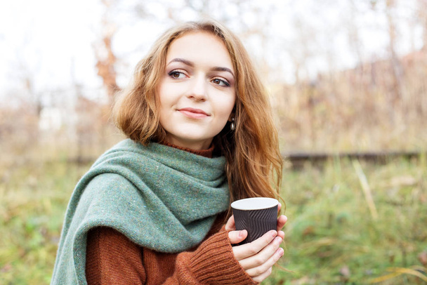 Young adult woman in a brown knitted sweater. Coffee in a plastic glass. The concept of a lifestyle, autumn, art, cosmetics and care. Nature. The concept of a lifestyle, autumn, cosmetics and care. - 写真・画像