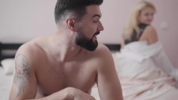 Portrait of young Caucasian bearded man looking back at satisfied blond woman sitting on bed at the background. Smiling guy resting in bedroom after sex. - Video