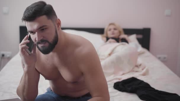Angry Caucasian bearded man talking on phone as sitting in bedroom. Young businessman leaving the room, his wife making desperate gesture at the background lying in bed. Overworking, hardworking. - Felvétel, videó