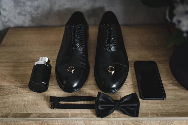 The groom is gathering in the morning. wooden shelf. Mens Black classic patent leather shoes. A black bow tie. Wedding details. Eau de toilette, black perfume, black mobile phone - Foto, Imagen