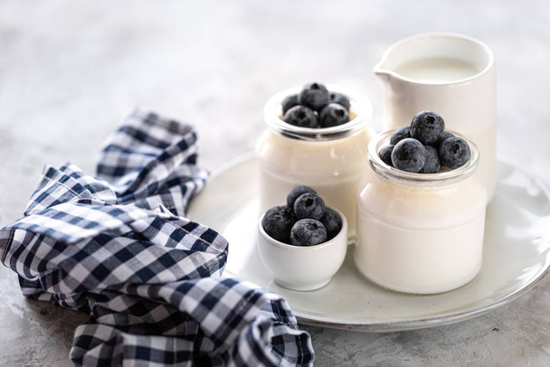 Melk en zelfgemaakte yoghurt in een pot op een wit bord. Slagroom met bosbessen als dessert in twee glazen potten. - Foto, afbeelding