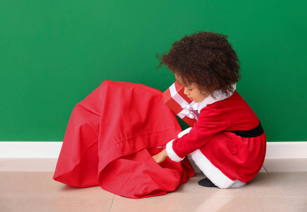 Little African-American girl in Santa costume and with bag full of gifts near color wall - Fotografie, Obrázek