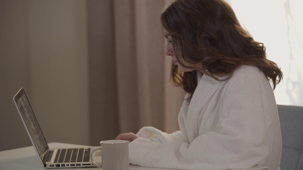 Side view close-up of charming Caucasian woman surfing Internet in the morning. Young brunette girl in white bathrobe sitting with laptop and coffee cup and smiling. Leisure, lifestyle, online. - Filmmaterial, Video