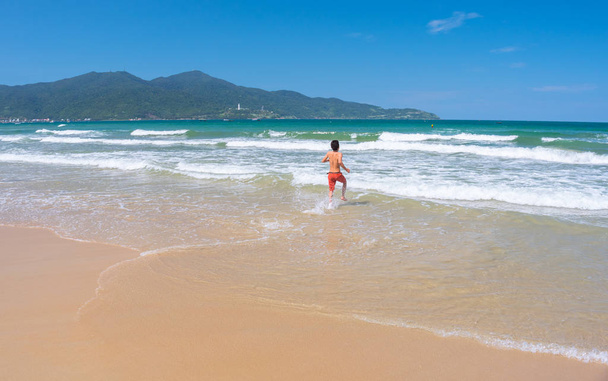 Da Nang Beach: an excited man runs through the surf to the gentle sea waves on My Khe Beach. Son Tra Peninsula and mountain on the horizon. This is the central beach of Danang City, Vietnam, in May. - Photo, Image